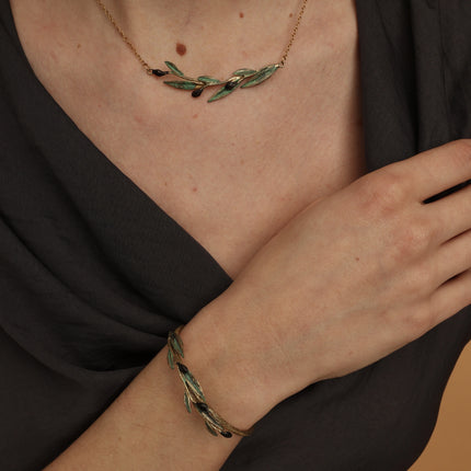 Handmade gold olive branch bracelet with green patina and black olive details, worn on a woman's wrist against a dark background