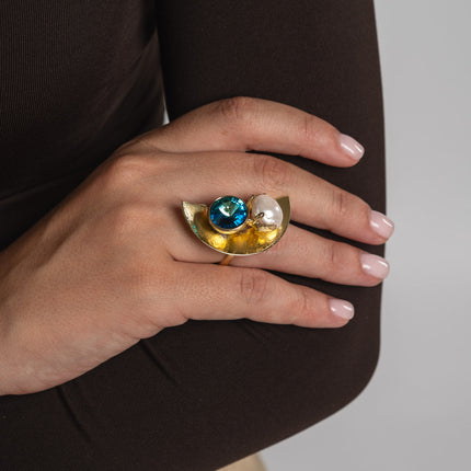 Artisan gold-tone brass architectural cocktail ring with blue crystal and baroque pearl on half-moon base worn on hand — Orbit Ring by Kalliope Jewelry, crafted in Greece