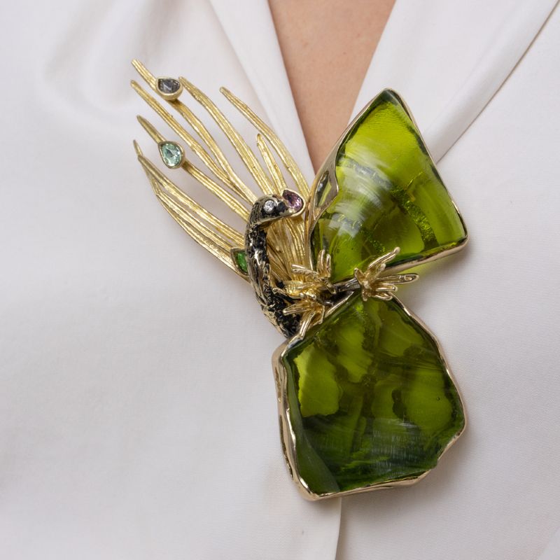 Woman wearing the Plume peacock brooch with large olive green resin stones, gold feathers and teardrop crystals on a white shirt, Savage Flora