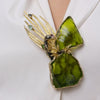 Woman wearing the Plume peacock brooch with large olive green resin stones, gold feathers and teardrop crystals on a white shirt, Savage Flora