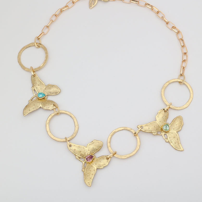 Flat lay of Nymia gold butterfly statement choker necklace with hammered ring links and faceted blue topaz, pink tourmaline and mint green crystals