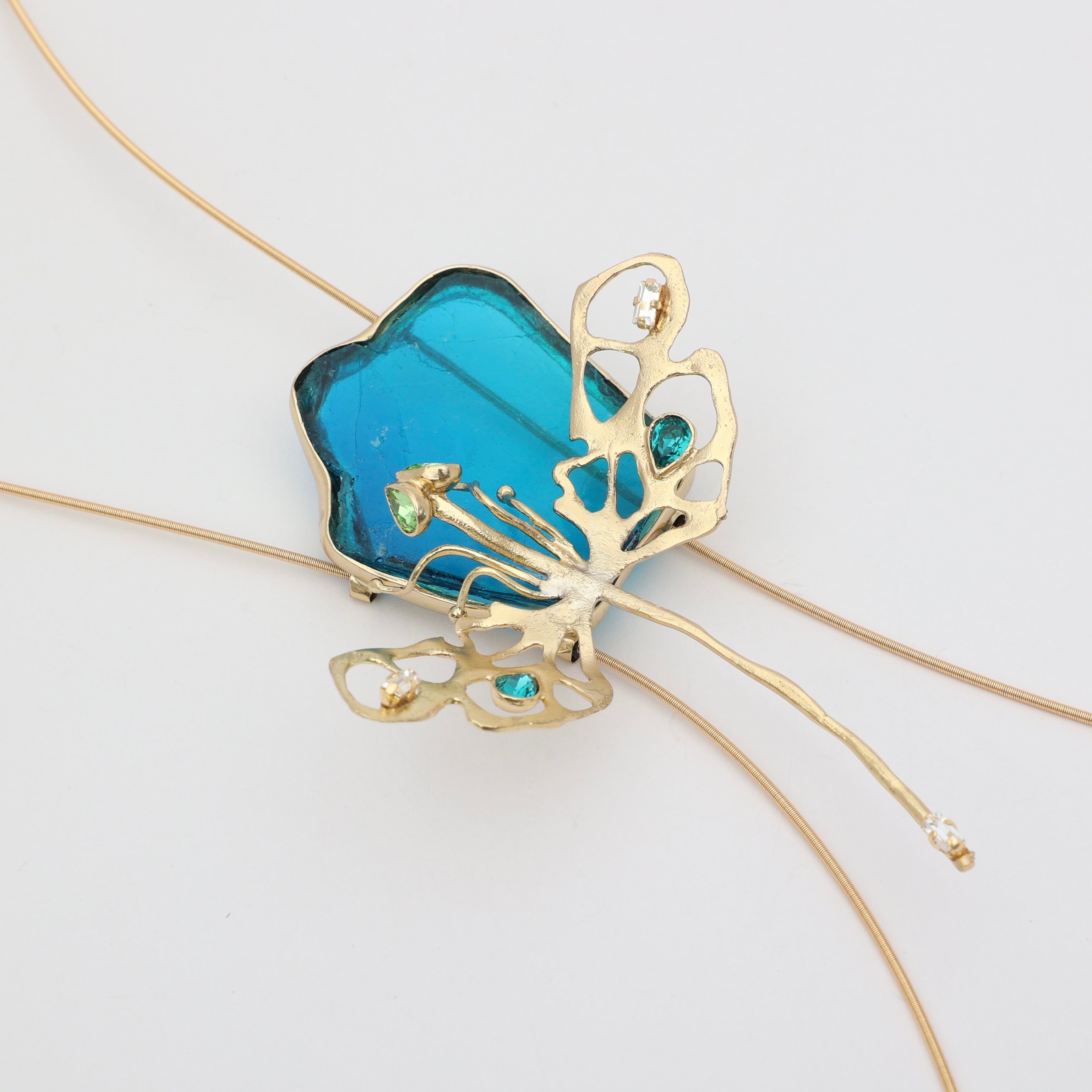 Close-up flat lay of Zephyra handmade brass necklace pendant with translucent blue resin flower, openwork gold leaves and teal crystals