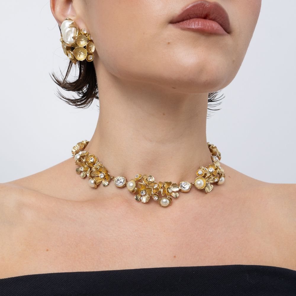 A close-up of a model wearing a gold-toned choker necklace and matching drop earrings decorated with gold, white pearl, and clear rhinestone elements against a light gray background.
