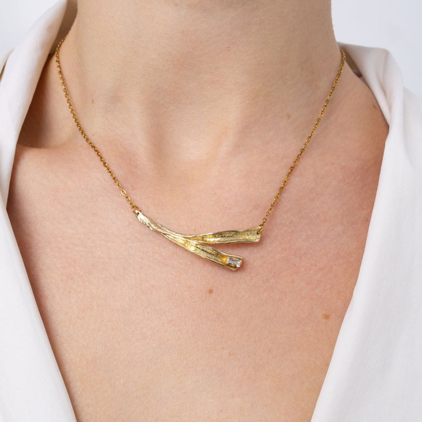 Woman wearing the Pixie gold-plated bar necklace with two crossed leaf-shaped pendants and a baguette crystal on a fine gold chain, Savage Flora