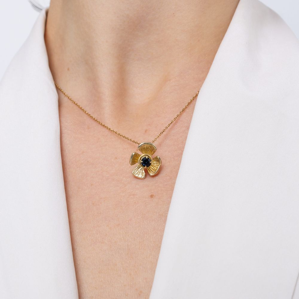 A gold pendant necklace featuring a stylized flower with a small dark blue-green center, worn on a model's neck over a white collared shirt.