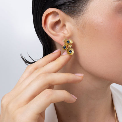 A close-up lifestyle shot of a model wearing a gold flower-shaped stud earring with small blue and yellow gemstones, with her hand gently touching her ear.