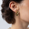 A close-up of a model's ear wearing a gold flower-shaped stud earring with three small purple gemstone accents.