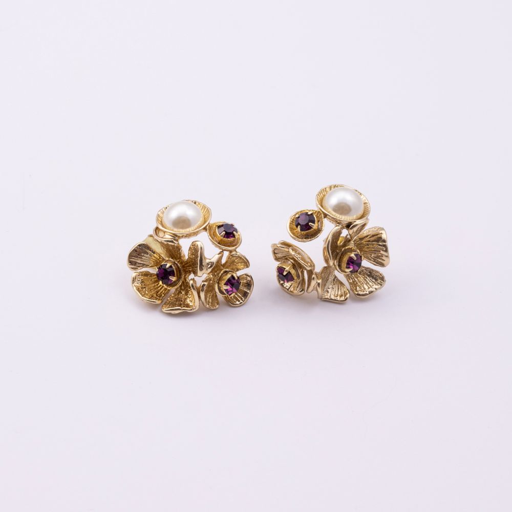 A pair of gold-toned flower-shaped stud earrings with white faux pearls and small dark purple gemstone accents, set against a plain white background.
