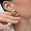 A close-up of a model's ear and hand wearing a gold flower-shaped stud earring with a central pearl and small clear rhinestones.