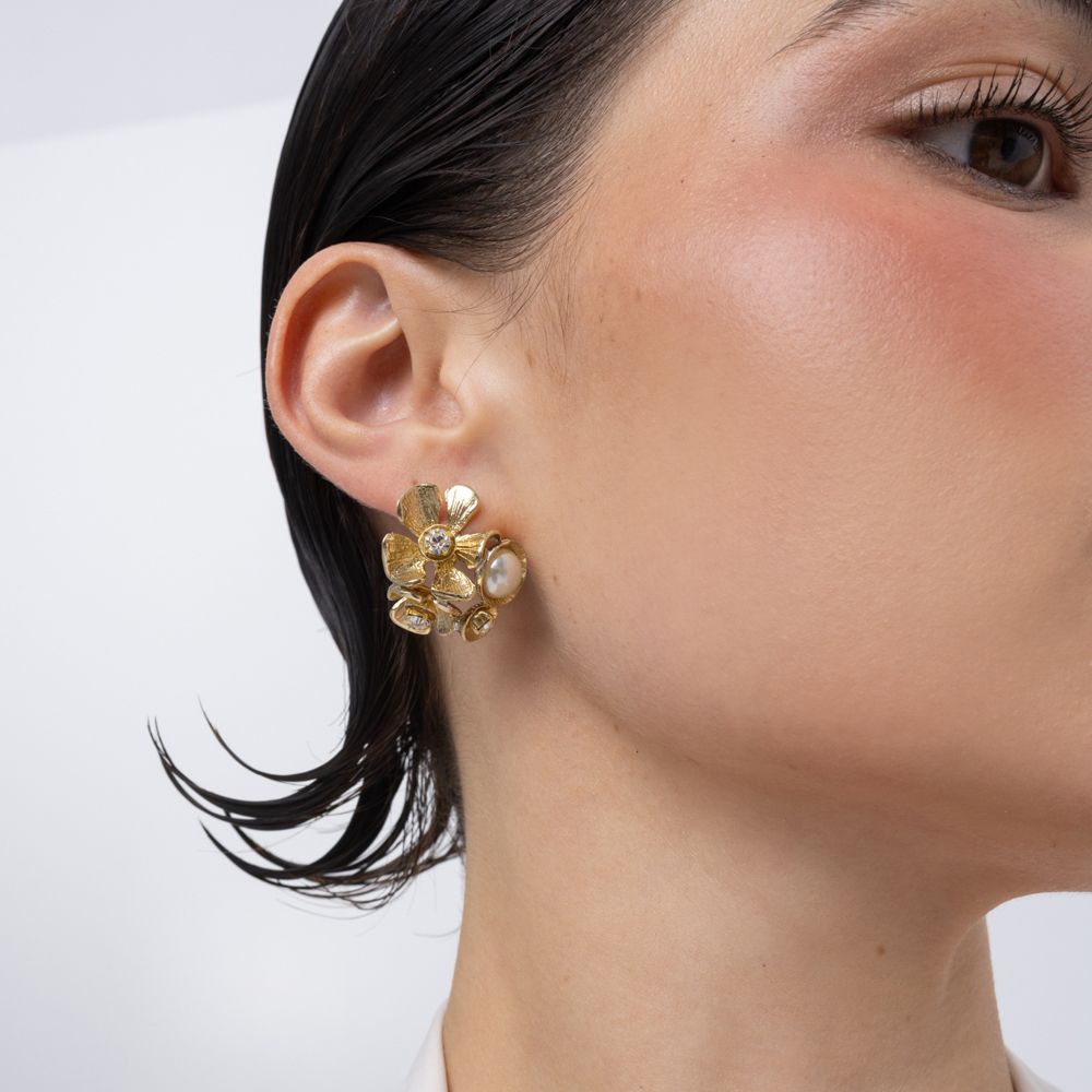 A close-up of a model's ear wearing a gold flower-shaped stud earring with clear rhinestones and a small pearl accent, against a plain white background.