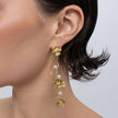 A close-up of a model's ear wearing a long, dangling gold earring with three gold flower charms, small white pearls, and clear rhinestones against a light gray background.