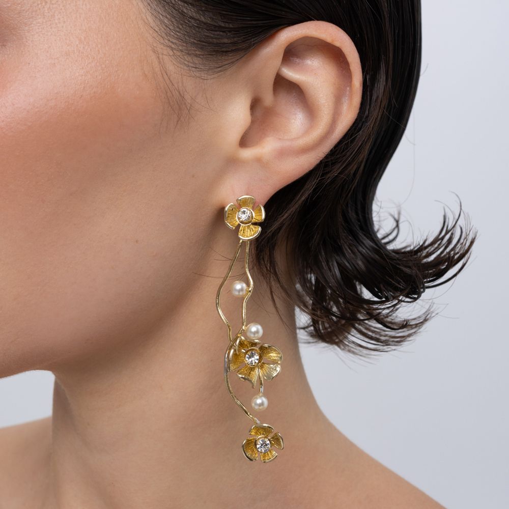 A close-up of a model's ear wearing a long, dangling gold earring with three gold flower charms, small white pearls, and clear rhinestones against a light gray background.