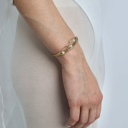 Handmade gold bangle bracelet with three white pearls and interwoven braided lines, worn on a woman's wrist against a white background
